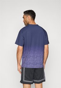 Αθλητικό μπλε ναυτικό μπλουζάκι με βαθμιαία μοβ σχέδια, κοντά μανίκια και στρογγυλή λαιμόκοψη, φορεμένο με γκρι σορτς από δίχτυ και λευκές λεπτομέρειες.