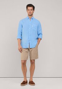 Camicia in lino azzurro chiaro con bottoni e taschino sul petto, abbinata a pantaloni corti beige e scarpe slip-on marroni, su uno sfondo neutro.