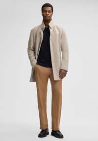 Beige wollen mix jas met een hoge kraag, marineblauw gebreid sweater eronder, gecombineerd met beige broeken en zwarte veterschoenen.