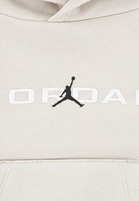 Beige hoodie med svart Jordan-logotyp och vit "JORDAN"-text. Tillverkad av mjukt tyg med en texturerad yta och en framficka.