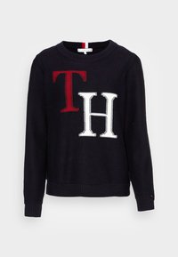 Zwarte gebreide trui met grote rode "T" en witte "H" initialen. Ronde halslijn, geribbelde manchetten en zoom met een contrasterende streepdetail.