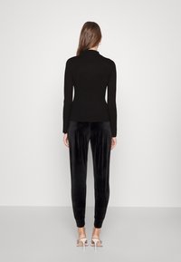 Schwarzes, figurbetontes, geripptes Oberteil mit langen Ärmeln, kombiniert mit schwarzen Samtjogginghosen und weißen, knöchelhohen, hochhackigen Sandalen. Neutraler Hintergrund.