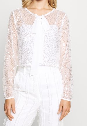 Bílá průsvitná halenka s dlouhým rukávem, flitry a zavazováním vpředu, v kombinaci s bílými kalhotami se svislými texturovanými pruhy.