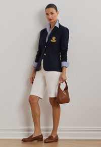Ναυτικό μπλέ σακάκι με χρυσά κουμπιά και κεντημένο λογότυπο, συνδυασμένο με ριγέ πουκάμισο, λευκά σορτς, καφέ μοκασίνια και καφέ τσάντα χειρός.
