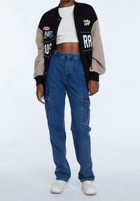 Chaqueta universitaria negra y beige con parches decorativos, combinada con pantalones cargo azules con bolsillos laterales y zapatillas blancas.
