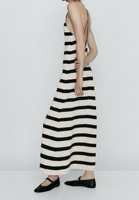 Robe maxi avec des rayures verticales noires et blanches, bretelles spaghetti, design dos nu et tissu texturé. Des ballerines noires portées avec la robe.