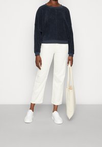Sudadera cortada de color azul marino, con una textura suave y mangas anchas, combinada con pantalones rectos blancos y zapatillas blancas. Bolsa tote beige.