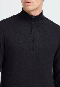 Pull en tricot noir avec un col à côtes haut et une fermeture éclair quart. Texture lisse et design ajusté, avec un léger éclat.