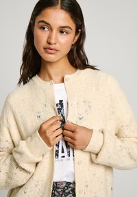 Cardigan a maglia crema con un motivo a diamante testurizzato, dotato di scollo rotondo e chiusura con bottoni, indossato sopra una T-shirt con grafica.