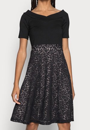 Femme portant une robe noire avec un décolleté bardot, des manches courtes et une jupe en dentelle fleurie longueur genou.