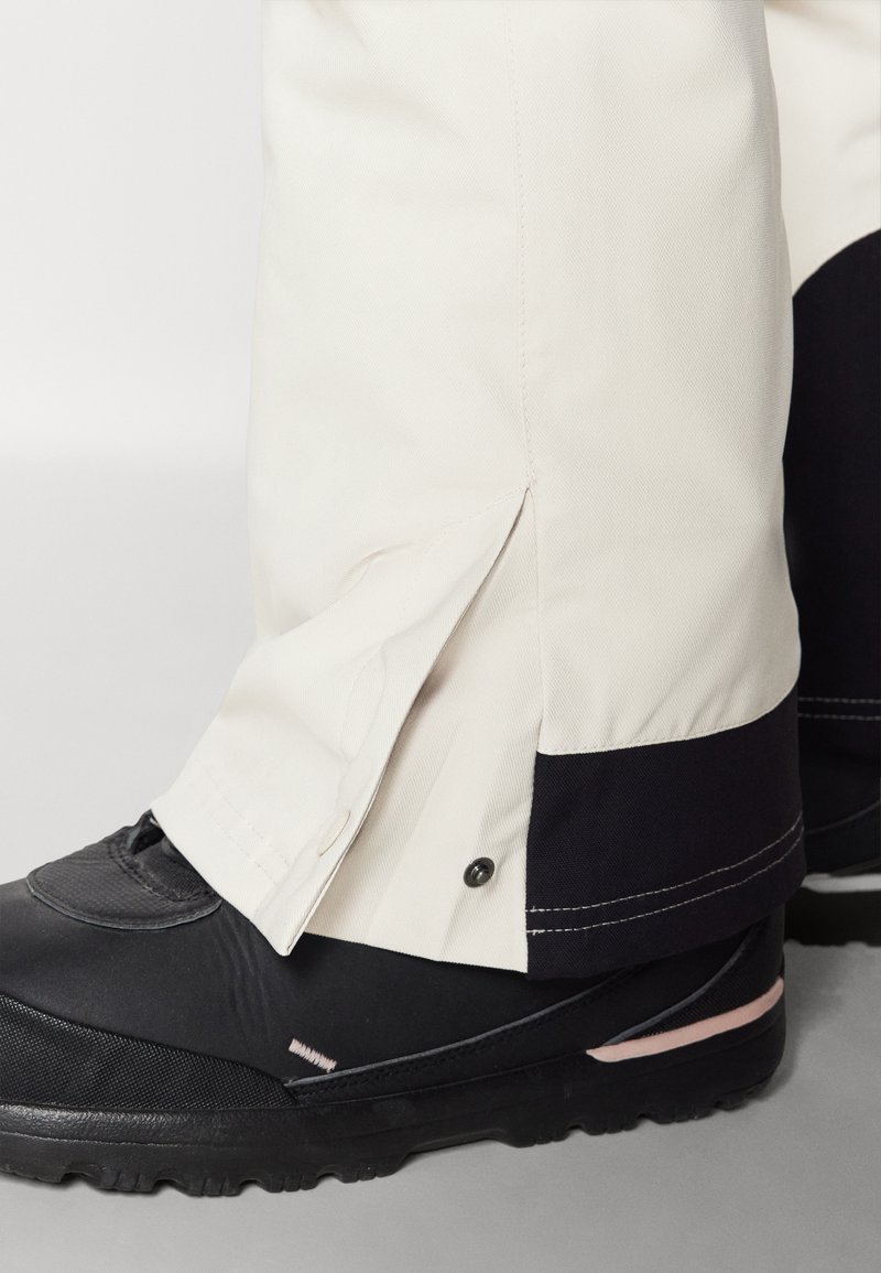 Biała nogawka spodni z czarnymi akcentami i zapięciem na zatrzask u dołu, w połączeniu z czarnym butem o teksturowanej powierzchni z różowym paskiem.
