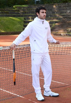 Bijela atletska trenirka s jaknom na zip i elastičnim manžetama, uparena s bijelim tenisicama s plavim i zelenim naglascima. Raketom za tenis u ruci.