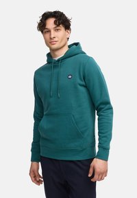 Sudadera de algodón color verde azulado con un bolsillo frontal, cordones ajustables y un pequeño logo en el pecho. Textura suave, corte casual y puños acanalados.