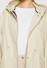Giacca da pioggia beige con zip frontale, cappuccio regolabile e tasche laterali. Presenta una fodera interna a righe e una vita regolabile con cordoncino per adattarsi meglio.