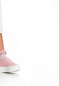 Laps, kes kannab roosakas platvorm-tossusid ja roosakaid volangiga sokke, seisab valge tausta ees.