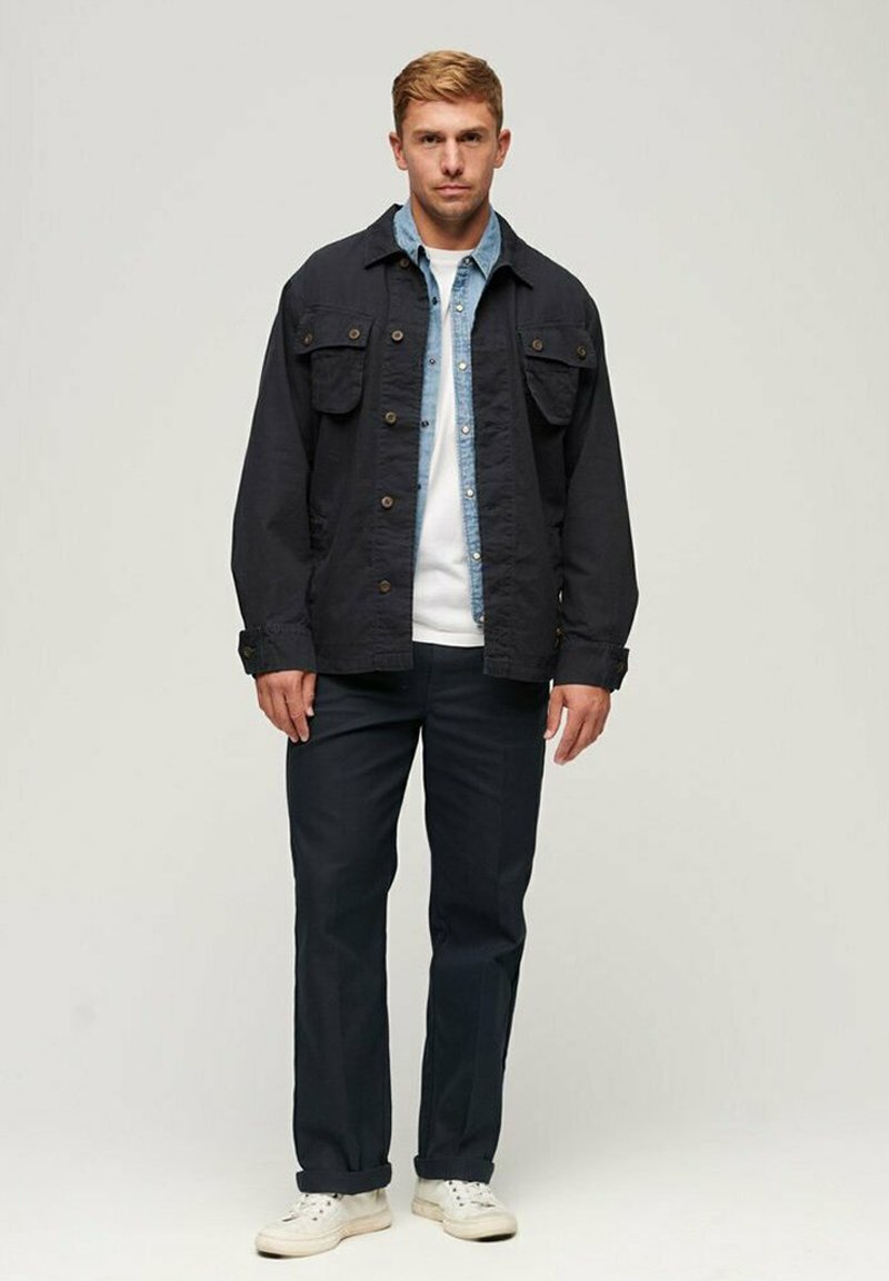 Joven de pie con chaqueta negra en capas sobre una camisa de mezclilla azul, pantalones negros y zapatillas blancas, contra un fondo liso.