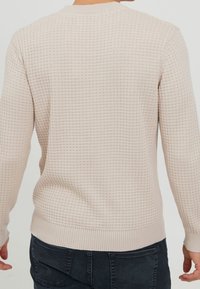 Pull en tricot beige clair avec une surface texturée et à motifs, col rond, ourlet et poignets côtelés. Porté avec un jean foncé.