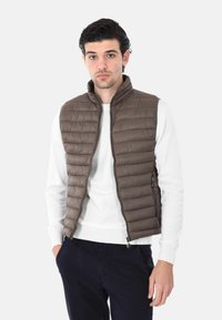 Gilet imbottito marrone con pannelli trapuntati orizzontali, colletto alto e zip frontale, indossato sopra un maglione bianco e pantaloni scuri.