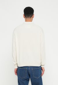 Moški s kratkimi kodrastimi lasmi, oblečen v belo dolgo majico z dolgimi rokavi in modre jeans hlače, obrnjena stran od preproste bele ozadja.