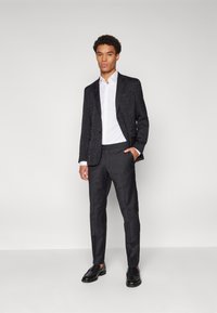 Schwarzer gemusterter Blazer über einem weißen Hemd, kombiniert mit dunklen Hosen und schwarzen Schuhen. Der Blazer hat ein strukturiertes Design und Revers.