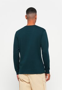 Man gezien van achteren in een donkere teal lange mouwen shirt en beige cargobroek tegen een effen witte achtergrond.