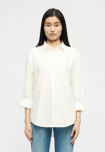 Νεαρή γυναίκα με μακριά σκούρα μαλλιά, φορώντας λευκό πουκάμισο με κουμπιά και μανίκια γυρισμένα, καθώς και μπλε τζιν, στέκεται μπροστά σε μια απλή φόντο.