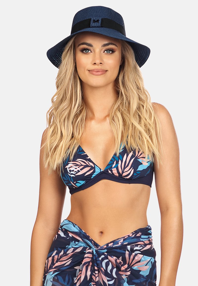 Kobieta z długimi blond włosami, nosząca niebieski kapelusz z szerokim rondem, kwiatowy staniczek bikini oraz pasującą spódniczkę na jasnoszarym tle.