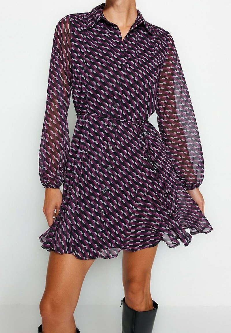 Femme portant une robe boutonnée violette à motifs avec des manches longues transparentes et des bottes noires mi-mollet, debout devant un fond blanc uni.