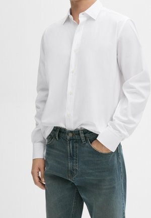 Person mit weißem langärmligem Hemd, das in eine blaue Jeans gesteckt ist, mit einer Hand in der Hosentasche, vor einfachem Hintergrund.