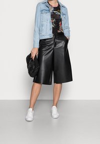 Kobieta w dżinsowej kurtce, kwiecistej bluzce, czarnych skórzanych culottes, białych sneakersach, trzymająca czarną puchową torebkę na tle jednolitego tła.