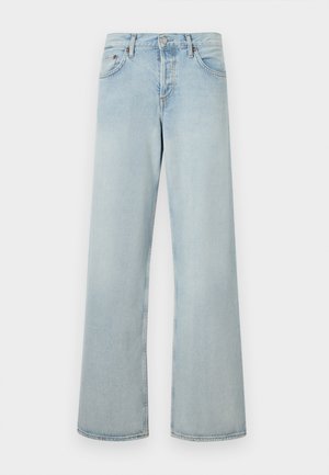 Femme debout portant un haut côtelé beige sans manches, un jean large bleu clair et des talons ouverts noirs sur un fond clair uni.