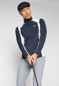 Navy and white zip-up golf jacket with long sleeves, featuring a high collar and sleek design. Grey pants and a matching navy cap complete the look.