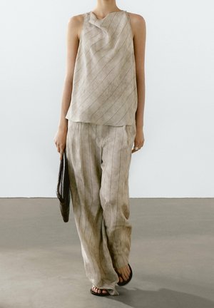 Kobieta ubrana w beżowy top bez rękawów i dopasowane szerokie spodnie z cienkimi pionowymi paskami, trzyma czarną kopertówkę i nosi czarne sandały.