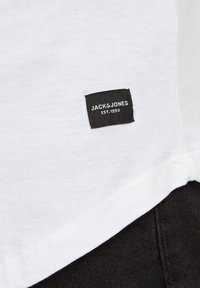 Witte katoenen t-shirt met een zwart geweven label aan de zoom, met de tekst "JACK & JONES EST. 1990." Soepele textuur en eenvoudig ontwerp.