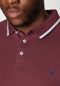 Burgundská polo košile s bílým pruhovaným límečkem, třemi knoflíkovými dírkami a vyšitým ptáčkem na levé hrudi. Texturovaná látka.