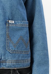 Kék farmerdzseki, mellzsebbel, amelyen egy fehér "Wrangler" címke található, narancssárga varrással és cikkcakk mintával a zseb dizájnján.