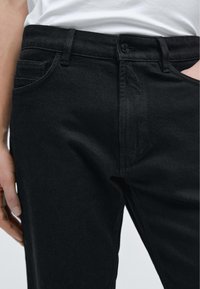 Černé džíny z denimu s rovnými nohavicemi, mají zapínání na knoflíky vpředu, boční kapsy a hladký, mírně texturovaný povrch.