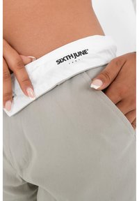 Lichtgrijze broek met een witte tailleband met een zwart logo. Glad materiaal, zijzak zichtbaar, persoon die de tailleband vasthoudt.