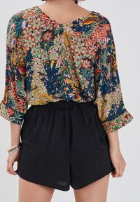 Bluzka w kwiatowy wzór z dekoltem w kształcie litery V, w żywych kolorach, w tym zielonym, czerwonym i różowym, połączona z czarnymi szortami oraz srebrnymi bransoletkami.