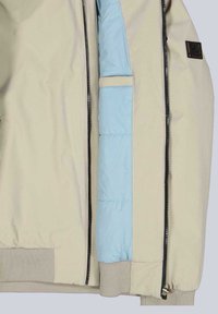 Hellbeige Jacke mit blauem Innenfutter. Verfügt über einen Reißverschluss vorne, elastische Bündchen und eine Tasche auf der linken Seite des Futters.