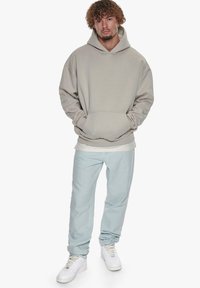 Lys grå oversized hoodie med en kengurulomme, struktureret stof og justerbar snørehette. Båret med lyseblå jeans og hvide sneakers.