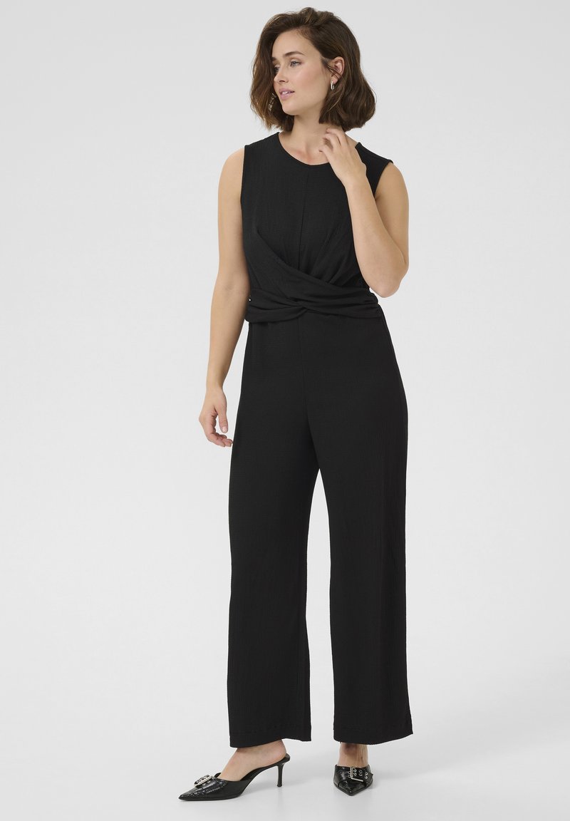Tuta nera con busto aderente e pantaloni larghi. Il tessuto presenta una sottile texture con un design intrecciato in vita. Indossata con mule neri con tacco.