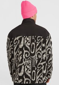 Veste floue noire et crème avec des motifs abstraits, présentant une partie supérieure noire uni et un bonnet rose côtelé porté par la personne.