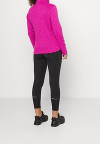Pullover in pile di colore rosa vivido con colletto alto, abbinato a leggings neri con dettagli riflettenti e scarpe da ginnastica nere.