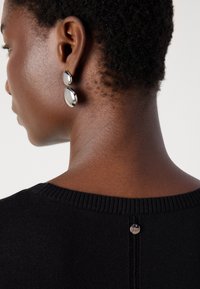 Black sweater from the back featuring a ribbed neckline, a small metal button at the center, and a textured neck area. Silver drop earrings visible.