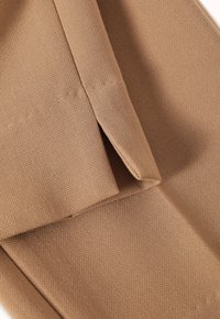Hnědá tkanina se strukturovaným tkaním, obsahující vrstvené záhyby. Materiál vypadá měkký a pružný, s teplým, tlumeným odstínem.