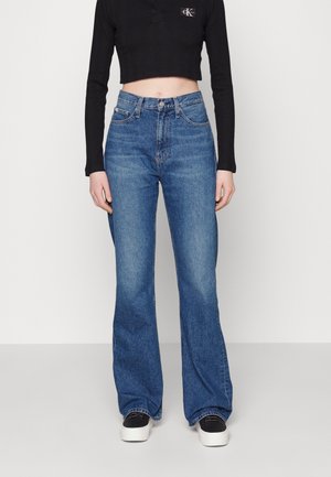 Kobieta w wysokich dżinsach z szerokimi nogawkami, czarnym krótkim topie z długim rękawem oraz czarno-białych sneakersach na platformie.