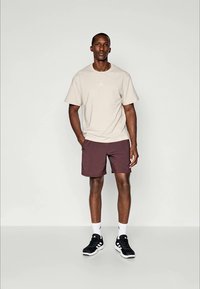 T-shirt beige avec un petit logo blanc, shorts bordeaux et baskets noires. Le modèle se tient sur un fond neutre avec des chaussettes blanches.