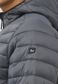 Sivka napihljiva jakna z quiltano teksturo, prilegajoča kapuca in majhna črna logotipna nalepka na ramu. Mehka, gladka tkanina.