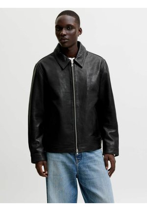 Osoba nosiaca čiernu koženú bundu na zips s golierom a svetlomodré rifle, stojaca pred jednoliatym bielym pozadím.